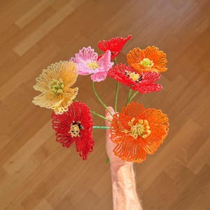 Puede incluir: Un ramo de flores de cuentas hechas a mano en tonos rojos, naranjas, rosas y amarillos. Las flores se sostienen sobre un fondo de madera. Las flores de cuentas son un artículo decorativo único y colorido.