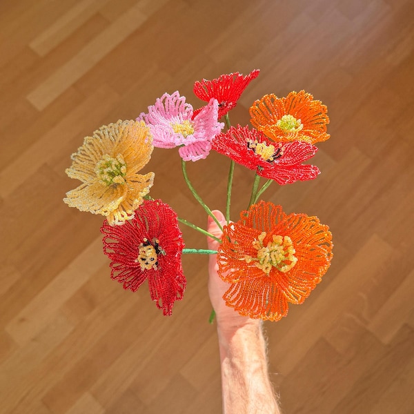Pode incluir: Um buquê de flores de contas feitas à mão em tons de vermelho, laranja, rosa e amarelo. As flores são seguradas contra um fundo de madeira. As flores de contas são um item decorativo único e colorido.