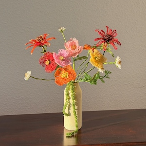 Może przedstawiać: Bukiet kolorowych kwiatów z koralików w kremowym wazonie. Kwiaty obejmują czerwone, pomarańczowe, różowe i żółte kwiaty z zielonymi łodygami. Wazon stoi na ciemnobrązowej drewnianej powierzchni.