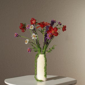 Könnte beinhalten: Eine cremefarbene Vase mit grünen Moosakzenten enthält einen bunten Blumenstrauß. Das Arrangement umfasst rote, violette, weiße und rosa Blüten. Die Vase steht auf einem weißen Tisch vor einem neutralen Hintergrund.