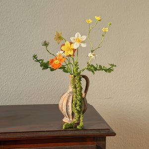 Könnte beinhalten: Ein Stillleben mit einer kleinen, beigefarbenen Vase mit Henkel, gefüllt mit bunten Perlenblumen. Die Blumen umfassen orange, gelbe und weiße Blüten mit grünen Stielen und Blättern. Die Vase steht auf einer dunklen Holzoberfläche.