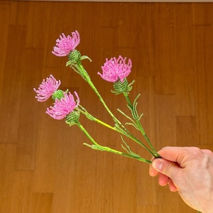 Puede incluir: Un ramo de flores de cuentas hechas a mano con flores de cardo rosas y tallos verdes. Las flores se sostienen contra un fondo de madera. Las flores de cuentas son un artículo decorativo único.