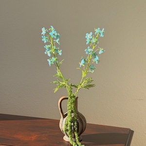 May include: A small, brown ceramic vase with a handle holds two stems of blue flowers and green foliage. The vase sits on a dark wooden surface, with a neutral-colored wall in the background. The arrangement is simple and elegant.