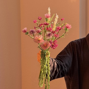 Puede incluir: Un ramo de flores de cuentas hechas a mano en tonos rosa, magenta y morado, con tallos de alambre intrincados y follaje de cuentas verdes. El arreglo incluye varias formas y texturas florales, todas construidas con pequeñas cuentas de vidrio.