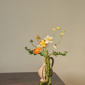 May include: A small, tan vase with a handle holds a bouquet of colourful, handmade flowers. The flowers include orange, yellow, and white blooms with green stems and leaves. The vase sits on a dark brown wooden surface against a neutral wall.