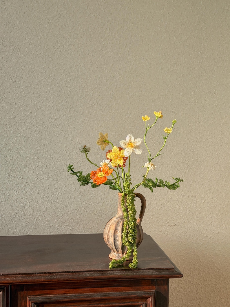 May include: A small, tan vase with a handle holds a bouquet of colourful, handmade flowers. The flowers include orange, yellow, and white blooms with green stems and leaves. The vase sits on a dark brown wooden surface against a neutral wall.