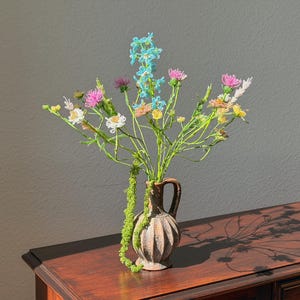 Op de afbeelding: Een boeket van kleurrijke kunstbloemen in een kleine, bruine, geribbelde vaas met een handvat. De bloemen zijn roze, blauw, geel en wit. De vaas staat op een donkere houten tafel, met de schaduw van de bloemen.