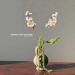 Puede incluir: Un peque&ntilde;o jarr&oacute;n r&uacute;stico contiene delicadas ramas florales blancas y rosas y enredaderas verdes. El jarr&oacute;n est&aacute; sobre una superficie de madera contra una pared gris neutra. El texto "DIGITAL PDF PATTERN" es visible.