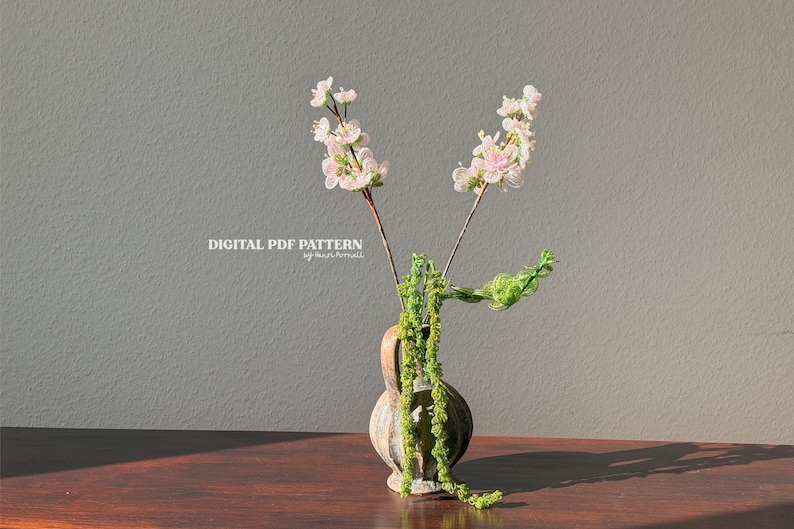 Puede incluir: Un peque&ntilde;o jarr&oacute;n r&uacute;stico contiene delicadas ramas florales blancas y rosas y enredaderas verdes. El jarr&oacute;n est&aacute; sobre una superficie de madera contra una pared gris neutra. El texto "DIGITAL PDF PATTERN" es visible.