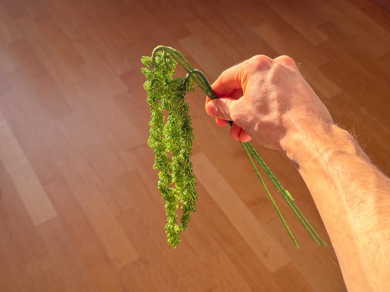 May include: A sprig of bright green artificial foliage with a cascading, textured appearance. The plant is held against a light brown wooden surface, with the sunlight highlighting the details of the leaves and stems.