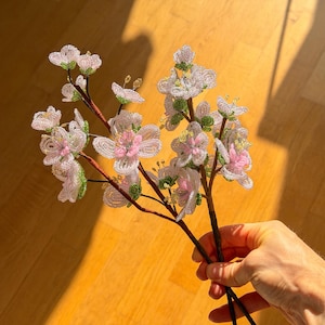 Puede incluir: Un arreglo floral de cuentas con delicadas flores blancas y rosas y detalles verdes. Las flores est&aacute;n unidas a tallos marrones, creando un aspecto naturalista. El arreglo se sostiene sobre un fondo c&aacute;lido de madera.