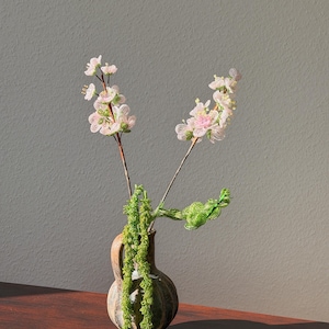 Könnte beinhalten: Eine kleine, rustikale Keramikvase enthält zarte Perlenblumen mit rosa und weißen Blüten. Grüne Perlenranken fallen kaskadenartig aus der Vase und schaffen eine natürliche, handgefertigte Ästhetik. Die Vase steht auf einer dunklen Holzoberfläche.