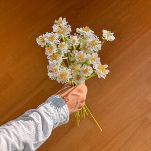 Könnte beinhalten: Eine Hand hält einen Strauß aus weißen und gelben Perlenblumen mit grünen Stielen. Die Blumen sind zusammengefasst, wobei die weißen Blütenblätter ein gelbes Zentrum umgeben. Der Hintergrund ist eine braune Holzoberfläche.