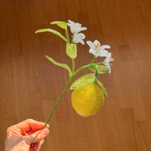 May include: A decorative botanical sculpture crafted from small glass beads, featuring a bright yellow lemon and two white lily flowers with yellow centres, all attached to a green wire stem with several textured green beaded leaves.