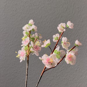 Puede incluir: Dos tallos de flores de cuentas con p&eacute;talos rosas y blancos, centros amarillos y detalles verdes. Las flores est&aacute;n sobre tallos marrones sobre un fondo gris. Las flores de cuentas son un art&iacute;culo decorativo.