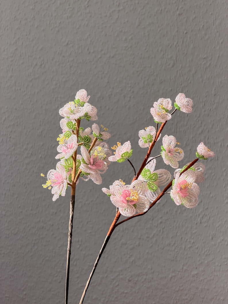 Puede incluir: Dos tallos de flores de cuentas con p&eacute;talos rosas y blancos, centros amarillos y detalles verdes. Las flores est&aacute;n sobre tallos marrones sobre un fondo gris. Las flores de cuentas son un art&iacute;culo decorativo.