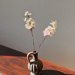Puede incluir: Un peque&ntilde;o jarr&oacute;n de cer&aacute;mica marr&oacute;n con asa contiene dos tallos de delicadas flores de cuentas rosas y blancas. El jarr&oacute;n descansa sobre una superficie de madera oscura, con una pared gris neutra en el fondo.