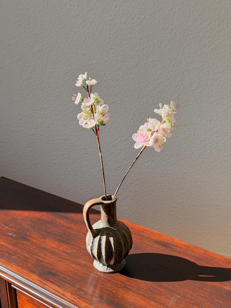 Puede incluir: Un peque&ntilde;o jarr&oacute;n de cer&aacute;mica marr&oacute;n con asa contiene dos tallos de delicadas flores de cuentas rosas y blancas. El jarr&oacute;n descansa sobre una superficie de madera oscura, con una pared gris neutra en el fondo.