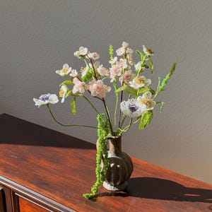 Puede incluir: Un peque&ntilde;o jarr&oacute;n marr&oacute;n contiene un ramo de flores hechas a mano. Las flores presentan flores blancas, rosas y moradas con tallos y hojas verdes. El jarr&oacute;n est&aacute; sobre una superficie de madera oscura, con sombras.