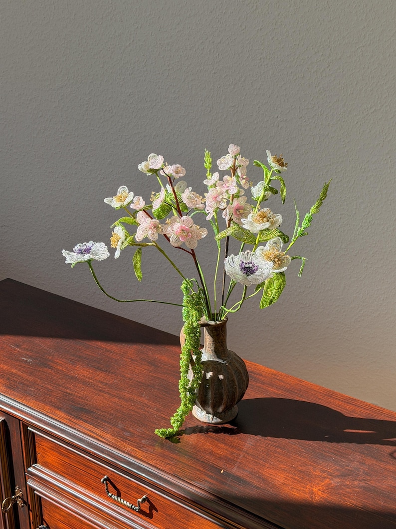 Puede incluir: Un peque&ntilde;o jarr&oacute;n marr&oacute;n contiene un ramo de flores hechas a mano. Las flores presentan flores blancas, rosas y moradas con tallos y hojas verdes. El jarr&oacute;n est&aacute; sobre una superficie de madera oscura, con sombras.