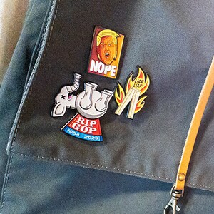 May include: Three enamel pins attached to a dark gray fabric surface. One pin features a portrait with the text NOPE. Another shows a white elephant with the text RIP GOP 1854-2020. The third pin depicts pants on fire with the text LIAR LIAR.