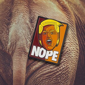 May include: A rectangular patch featuring a stylized illustration of a person with blonde hair and an orange complexion, mouth open in an expression of shouting. The word NOPE is printed in large white capital letters at the bottom of the patch.