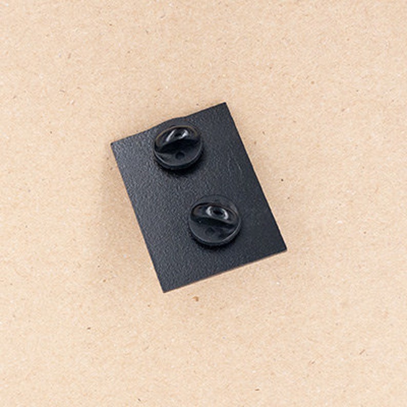 May include: A rectangular black metal pin backing featuring a textured surface and two circular black rubber clutch fasteners attached to the rear side. The pin back is designed to secure a decorative enamel pin to clothing or fabric accessories.
