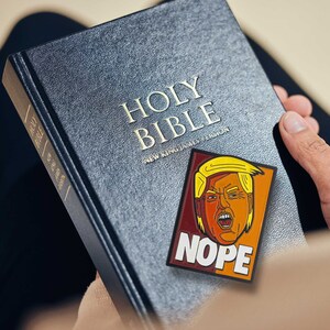 May include: A black Holy Bible New King James Version book featuring a rectangular patch with a yellow-haired figure and the text NOPE in white letters against a maroon and orange background, displayed on the front cover of the textured black book.