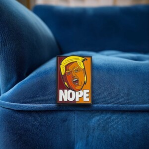 May include: A rectangular pin featuring a stylized illustration of a person with blonde hair and an orange complexion, appearing to shout with an open mouth. The background is split between dark red and orange, with the word NOPE in white letters.