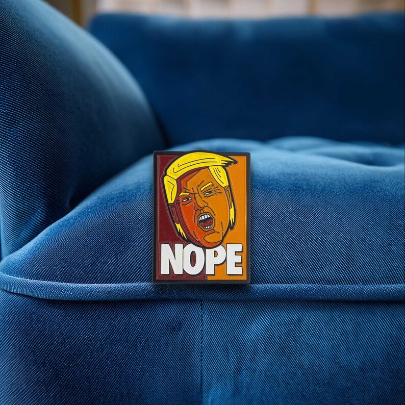 May include: A rectangular pin featuring a stylized illustration of a person with blonde hair and an orange complexion, appearing to shout with an open mouth. The background is split between dark red and orange, with the word NOPE in white letters.
