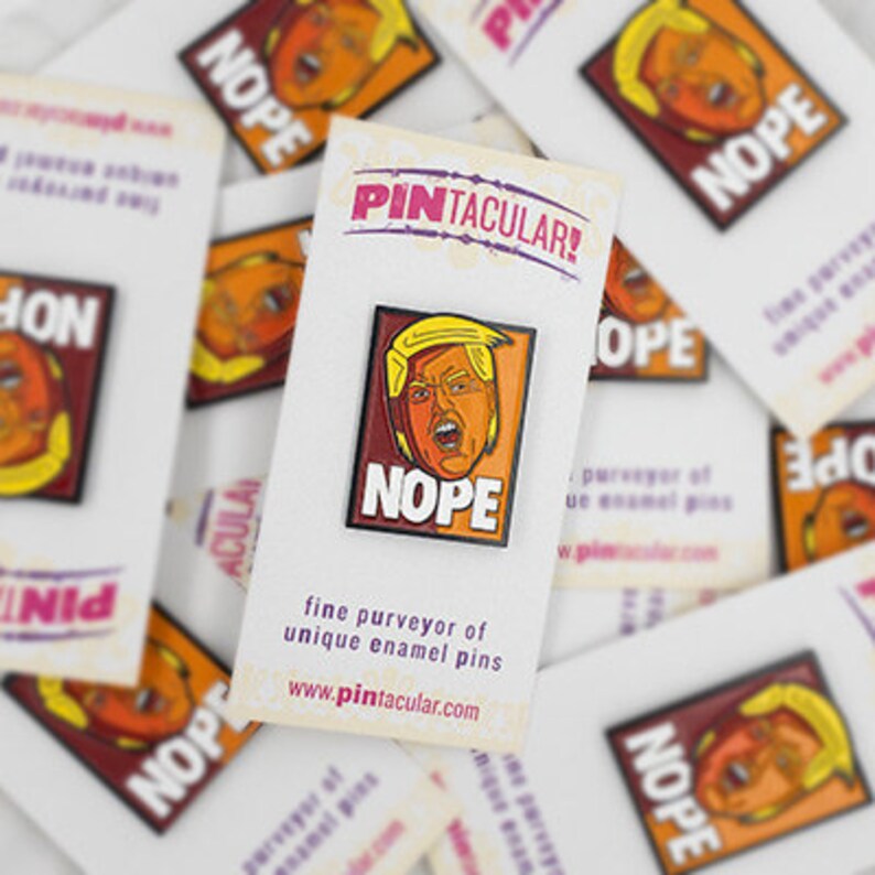 May include: A rectangular enamel pin featuring a stylized illustration of a person with blonde hair and an open mouth, set against a dark red and orange background with the word NOPE in white capital letters, mounted on a white display card.