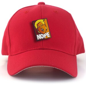May include: A bright red baseball cap featuring a rectangular enamel pin attached to the front. The pin displays a cartoon illustration of a person with blonde hair and the word NOPE printed in bold white capital letters on a dark background.