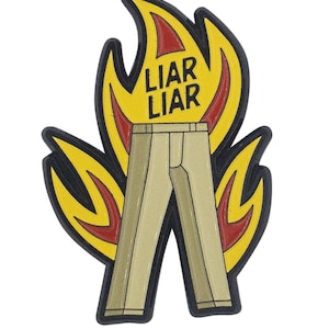 May include: A metal enamel pin featuring a pair of light tan trousers engulfed in bright yellow and red flames. The text LIAR LIAR is printed in bold black capital letters across the upper portion of the flames, centered above the trousers.