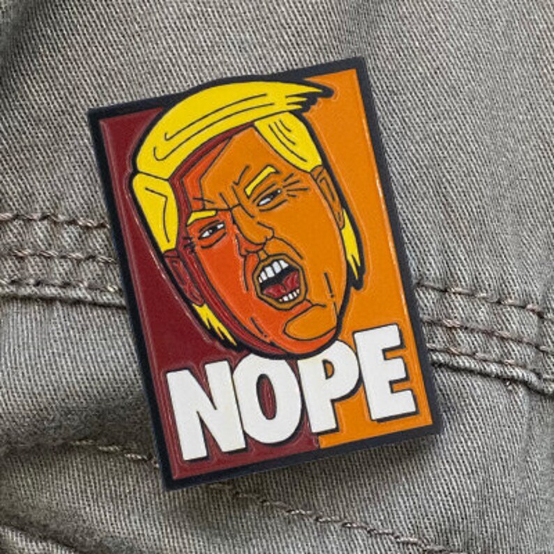 May include: A rectangular enamel pin featuring a stylized illustration of a person with yellow hair and an orange face, depicted with an open mouth. The background is split between dark red and orange, with the word NOPE in large white letters.