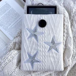 May include: White knitted tablet sleeve with three embroidered gray stars and a black button closure. The sleeve is resting on a cream-colored knitted blanket, with an open book in the background.