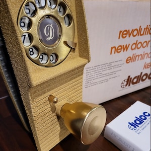 May include: A gold-colored Dialoc door lock with a rotary dial and a matching doorknob. The lock has a textured surface and a white box with the text "Dialoc" and "2-3/4" BACKSET 3". The box is next to the lock.
