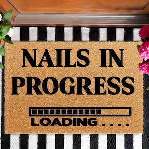 May include: A rectangular doormat with the words "NAILS IN PROGRESS" in black, with a loading bar graphic and the word "LOADING..." below. The mat is a light brown color and sits on a black and white striped surface.