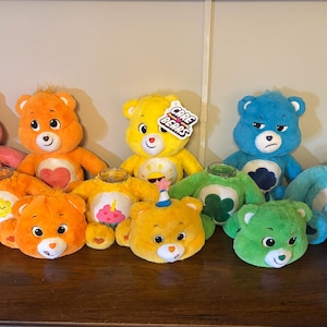 May include: A collection of Care Bears plush toys and jar-style containers featuring various colors including pink, orange, yellow, blue, and green. The items display embroidered belly badges such as a rainbow, sun, cupcake, cloud, and clover, with one yellow bear holding a Care Bears tag.