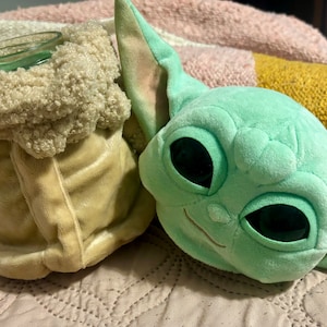 Pode incluir: Um brinquedo de pelúcia verde Baby Yoda com olhos grandes e uma pequena boca, deitado em um cobertor rosa e amarelo. O brinquedo está ao lado de uma caneca de cerâmica bege com uma borda de pele branca.