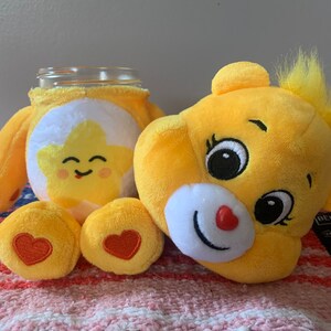 May include: A yellow plush Care Bear-themed jar cover and a Care Bear plush head. The jar cover features a smiling star design. The plush head has a red heart nose and a tuft of yellow hair.