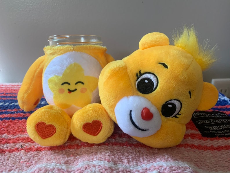 May include: A yellow plush Care Bear-themed jar cover and a Care Bear plush head. The jar cover features a smiling star design. The plush head has a red heart nose and a tuft of yellow hair.