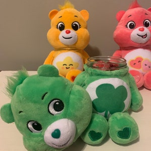 Puede incluir: Tres osos de peluche y un frasco de vidrio. Un oso amarillo con un emblema de estrella, un oso rosa con dos corazones y un oso verde con un emblema de trébol. La cabeza del oso verde está separada, revelando un frasco lleno de dulces rojos.