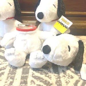 Puede incluir: Muñecos de peluche blancos de Snoopy con orejas y narices negras, collares rojos y una etiqueta amarilla que dice "PEANUTS". Un muñeco tiene un tarro de cristal con un borde rojo en lugar de su cabeza.