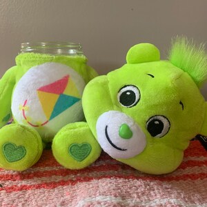 May include: A lime green plush bear with a jar body, featuring a white belly with a kite design. The bear has a smiling face, green heart-shaped accents, and a fluffy green tuft. The jar is clear glass.