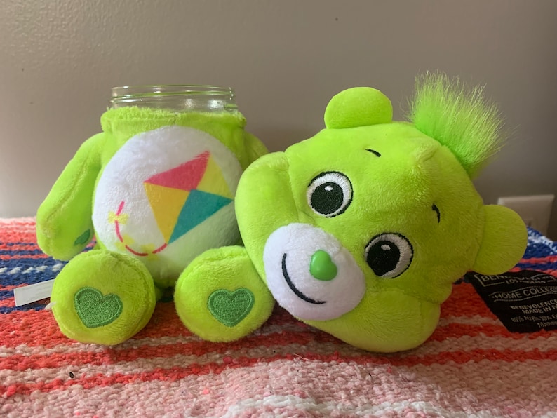 May include: A lime green plush bear with a jar body, featuring a white belly with a kite design. The bear has a smiling face, green heart-shaped accents, and a fluffy green tuft. The jar is clear glass.