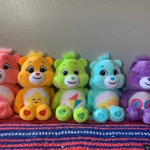 May include: Five plush Care Bears in various colors: pink, yellow, green, blue, and purple. Each bear has a unique belly badge and a heart on its foot. They are arranged in a row, showcasing their different designs.