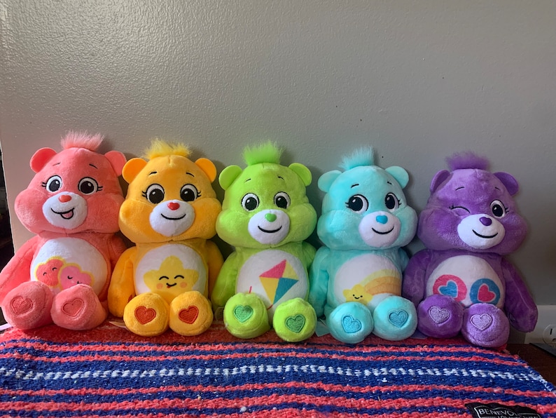 May include: Five plush Care Bears in various colors: pink, yellow, green, blue, and purple. Each bear has a unique belly badge and a heart on its foot. They are arranged in a row, showcasing their different designs.