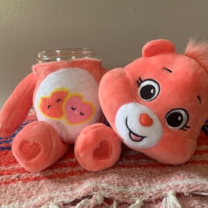 May include: A plush, coral-colored Care Bear-themed jar cover and matching plush bear. The jar cover features a white belly with two heart graphics. The bear has a heart-shaped nose and a tuft of hair. The bear's foot has a heart.