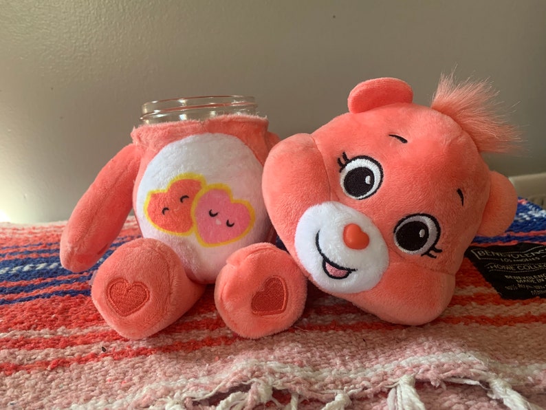 May include: A plush, coral-colored Care Bear-themed jar cover and matching plush bear. The jar cover features a white belly with two heart graphics. The bear has a heart-shaped nose and a tuft of hair. The bear's foot has a heart.