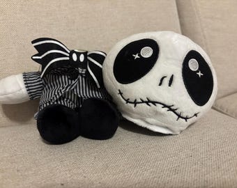 May include: A plush toy featuring a round white head with large black eye sockets and a stitched mouth, positioned next to a small body wearing a black and white pinstriped suit jacket, black trousers, and a large black and white striped bow tie.